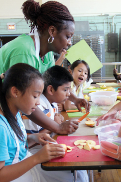 Cooking class: Haley House employee Vanessa Labranche assists Jonathan Delacruz (center) during the Dever summer program’s nutritional cooking class last Wednesday.  Nhu Huynh Duong (left) chops apples as Tiffany Nguyen (right) observes. Elizabeth Murray photo