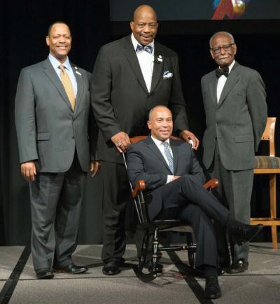 Former governor Deval Patrick tries out a new chair given to him by UMass Boston Chancellor J. Keith Motley (center) as a token of the university establishing an academic faculty chair in his honor. Also pictured are UMass Board of Trustees Chairman Victor Woolridge, left,  and UMass Boston Provost Winston Langley.