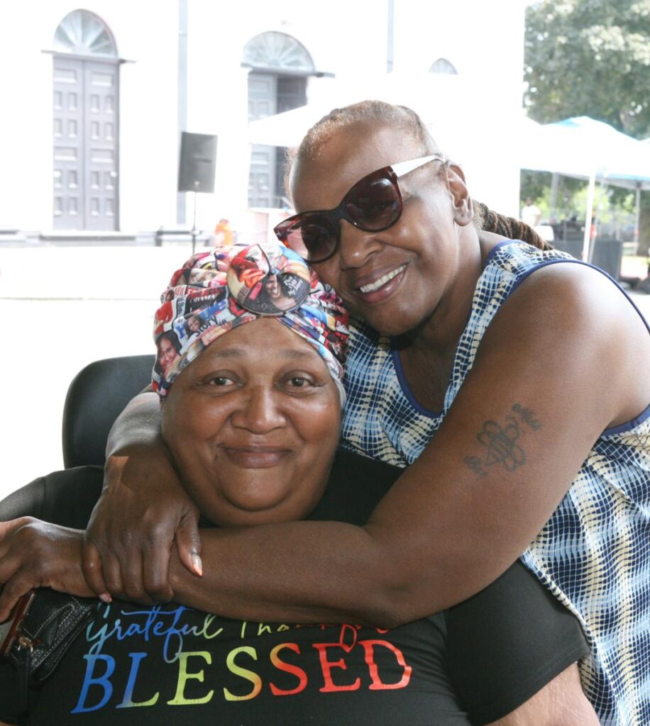 A woman, standing, hugs another woman, sitting. Both look at the camera smiling.