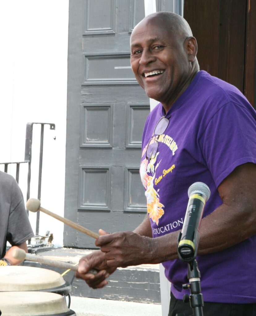 A man in a purple shirt plays the drum, smiling at the camera.