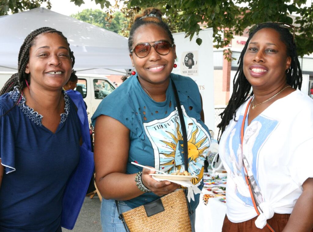 Three women eating jerk chicken, oxtail, and mac & cheese, smiling and looking at the camera.